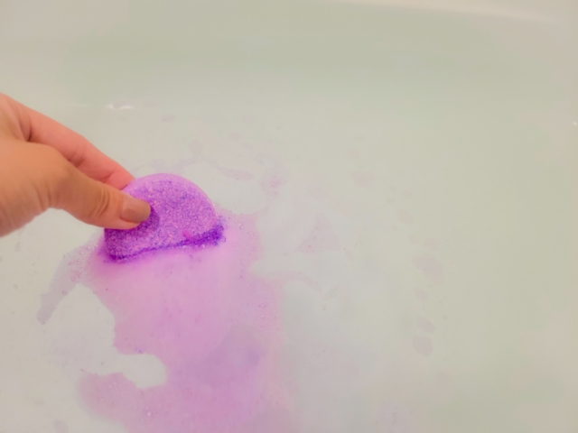 お湯をはった湯船に紫色の入浴剤を浸して溶かしている女性の手先