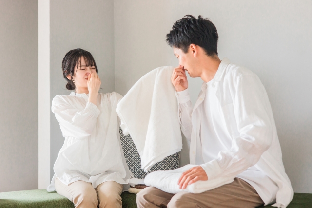 白いタオルを顔に近づけ洗濯物の生乾き臭に困る夫婦
