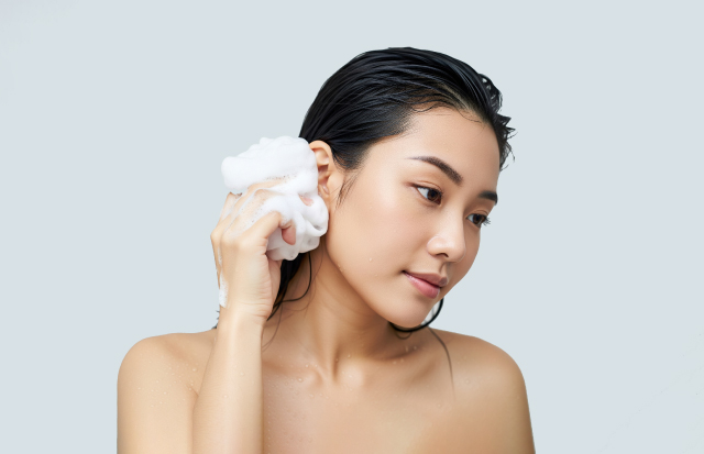 バスルームで泡がついたスポンジで耳の後ろを洗っている若い女性